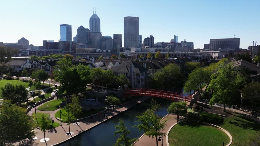 Orange pedestrian bridge crosses the Indianapolis Central Canal walk with downtown skyline in the background, aerial static