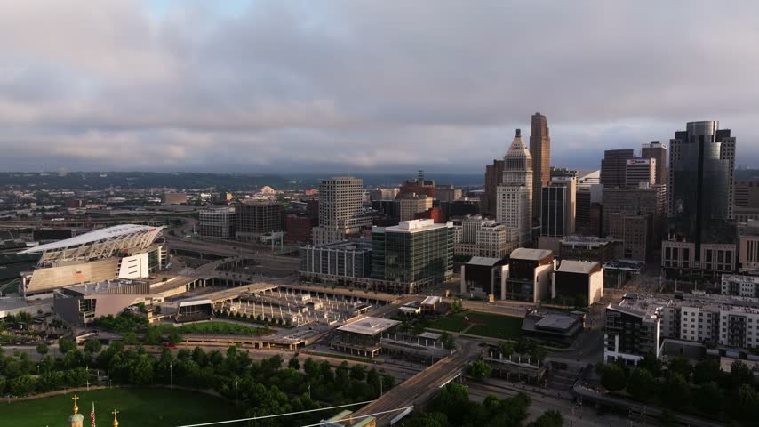 Downtown towers and civic buildings fill the Cincinnati riverfront beyond a historic suspension bridge, aerial pullback reveal