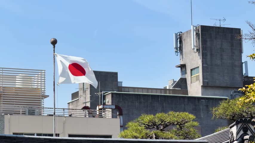 Japanese national flag blowing in breeze atop metal pole overlooking city architecture during sunny spring day cinematic footage.