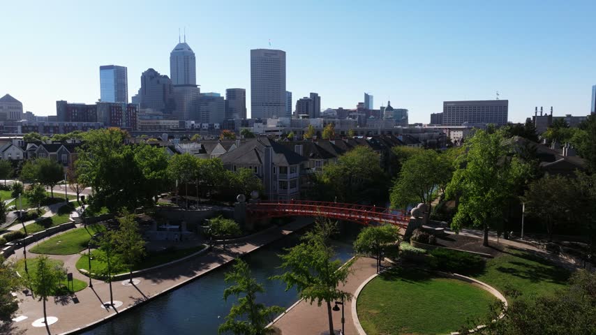 Downtown Indianapolis overlooks the Central Canal, landscaped paths and an orange pedestrian bridge, aerial dolly above recreational canal walk park