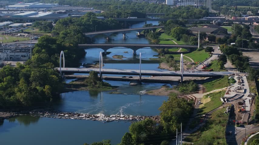 New modern bridge with large white arches spans an urban river beside older crossings and construction areas, aerial medium approach