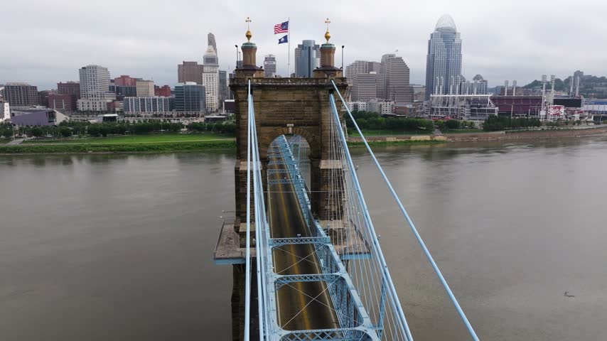 Roebling Bridge tower and blue suspension cables lead toward the downtown Cincinnati skyline across the Ohio River, aerial dolly ascend flyover