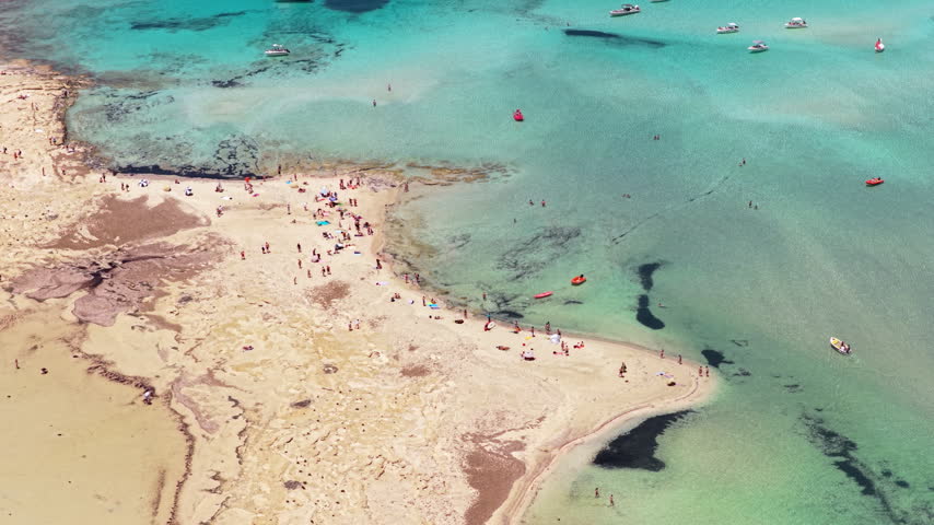 Aerial drone view of Balos Lagoon with boats offshore and sunbathers along the white sandbar, Balos, Greece