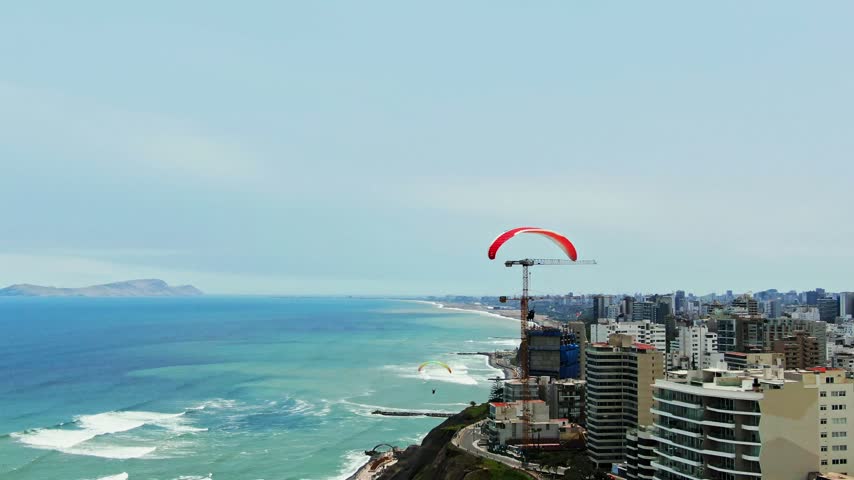 Drone follows a paraglider over the Miraflores coast in Lima, Peru, ending with three paragliders in the sky above the ocean, cliffs, cranes and seaside buildings.