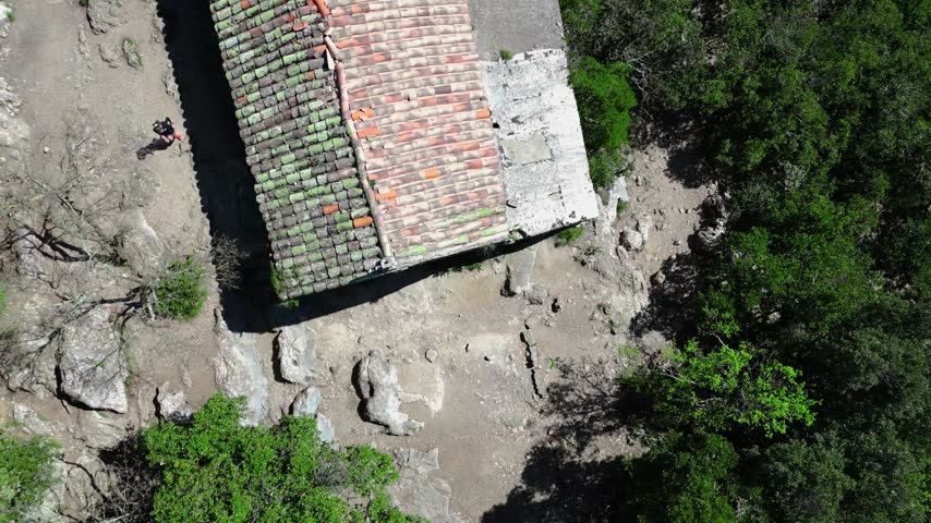A drone shot from above showing a section of forest on the right, with the roof of a chapel surrounded by a rocky path in full sunlight, and a person running