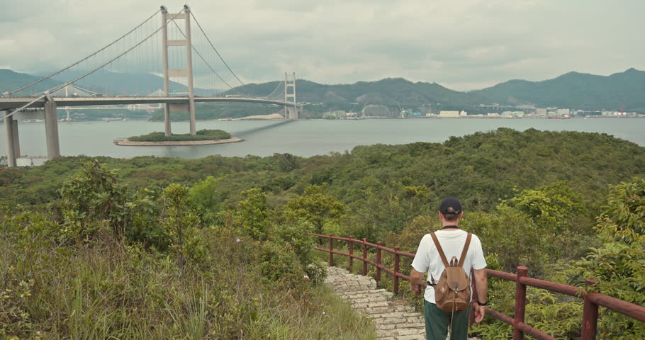 Man with backpack walking down stone stairs on hill, big Bridge background, Hong Kong landscape, outdoor travel and adventure tourism.
