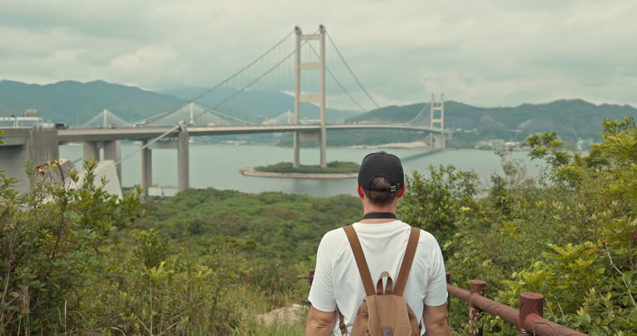 Traveler with backpack looking at bridge. Man on hill observing suspension bridge over water, travel and tourism lifestyle.