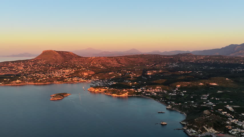Aerial drone view of a flat-topped hill above a coastal village and calm bay at golden hour, Chania, Greece