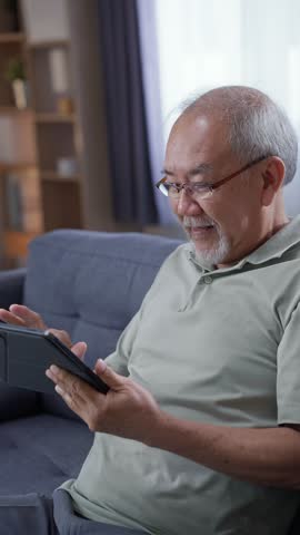 Smiling Asian older man using digital tablet computer sitting on sofa in living room during leisure time at home, technology and wireless communication