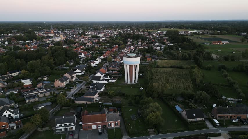 A water tower with surrounding buildings and green fields in Putte, Belgium, illuminated by soft evening light.
