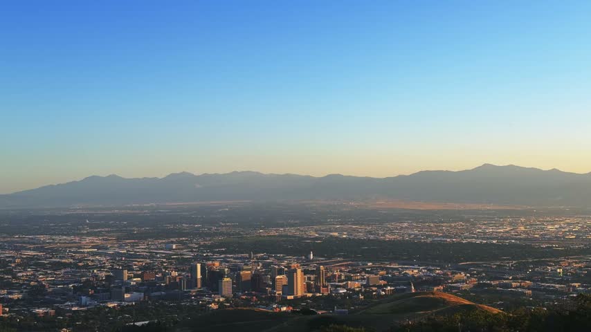 Tilting down wide shot of Salt Lake City and the valley from a hike on the Bonneville Shoreline Trail in Utah during golden hour on a warm spring evening, with soft light washing over the city below.