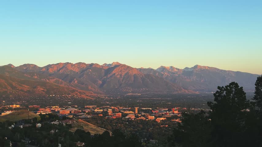 Tilting down wide shot of Salt Lake City and the valley from a hike on the Bonneville Shoreline Trail in Utah during golden hour on a warm spring evening, with soft light washing over the city below.