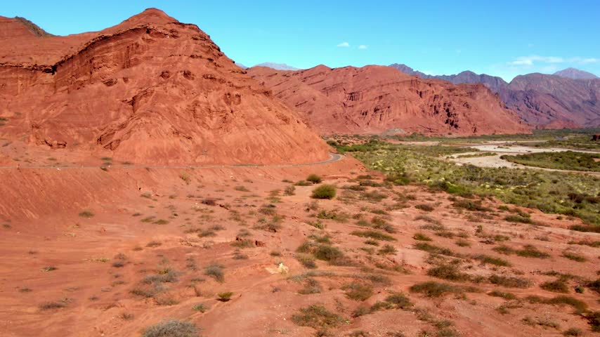 A narrow road curves around a massive red hillside above sparse scrub in Quebrada de las Conchas, a Salta tourist attraction. The elevated camera slowly pans across the sunlit landscape.