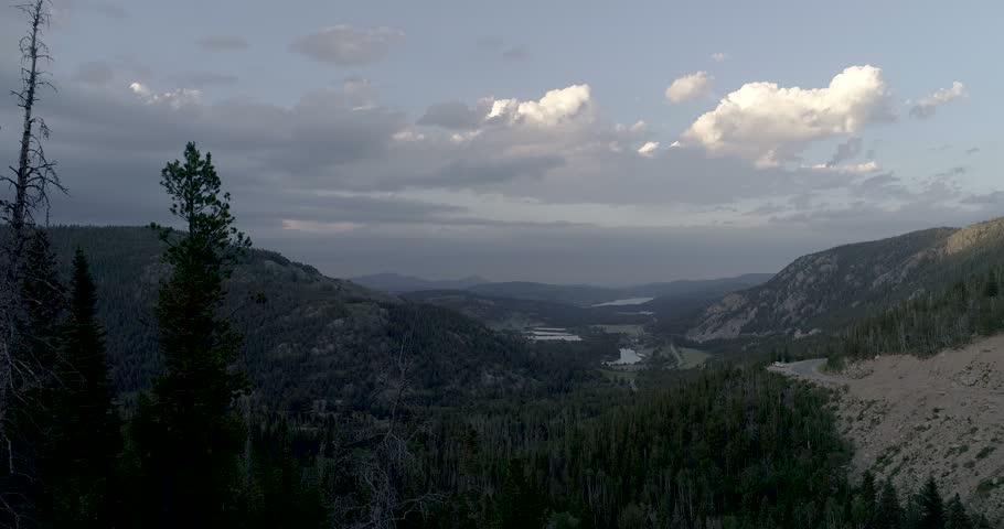 Scenic mountain landscape featuring dense evergreen forests winding asphalt highway and distant lakes under cloudy sky.