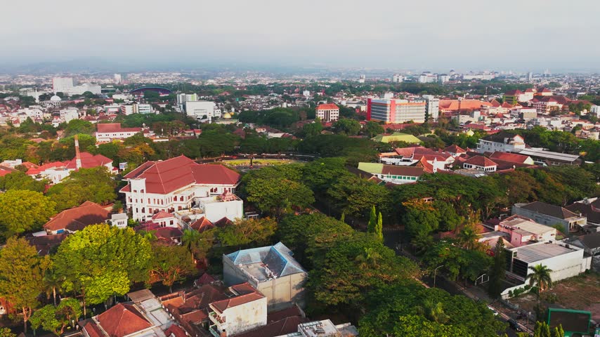 Aerial view of Malang City Hall surrounded by lush trees and historic red-roofed buildings in the urban landscape of Kecamatan Klojen, Jawa Timur, Indonesia.