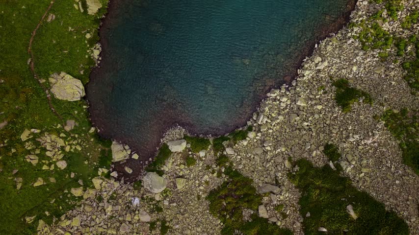 An aerial view pans upward over a dark mountain lake. The natural landscape features rocky shores and green grassy terrain. The top-down perspective captures water and land textures.