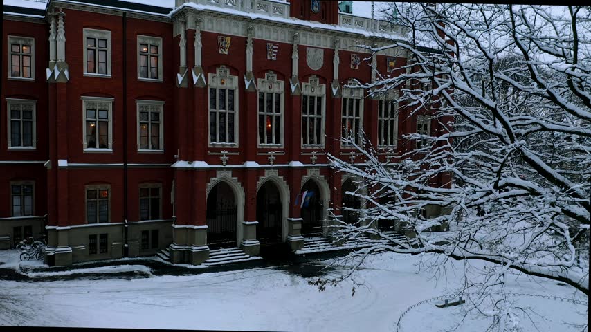 Rising drone shot showing iconic red brick university architecture with snowy roof and Wawel Castle in distance during overcast winter day.