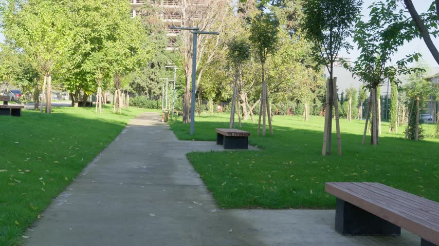 A paved park path passes wooden benches and a litter bin beneath leafy trees, with green lawns surrounding the quiet urban walkway in daylight. Tbilisi