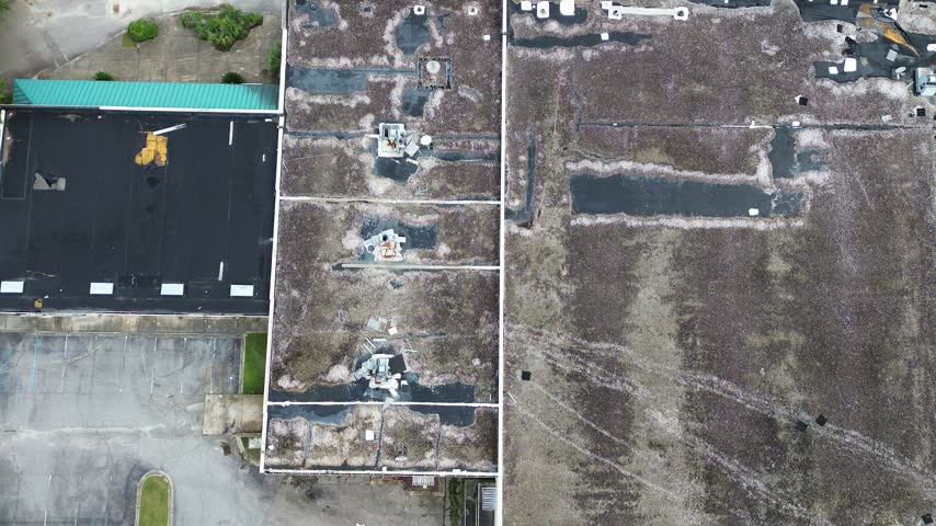 Tarnished ventilation equipment stands across patched gravel roofing of obsolete retail properties along Charleston. Wild grasses emerge through asphalt fractures alongside, South Carolina. USA.