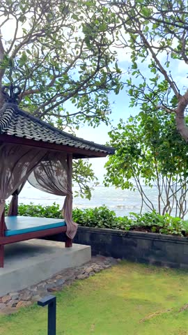 Seaside gazebo stands beside quiet lawn, framed by leafy trees and low stone wall. Open timber pavilion overlooks calm water, creating peaceful tropical garden retreat in daylight.