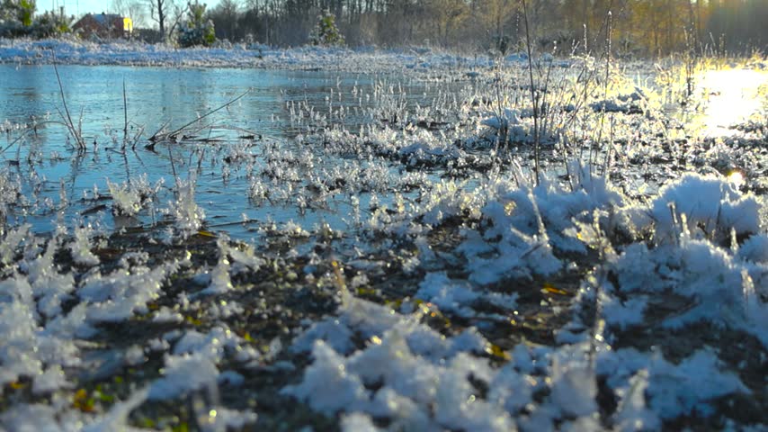 Macro view of frozen hoarfrost crystals on river bank. Beautiful winter landscape with morning sun and icy water.