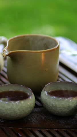 Steaming teapot rests beside two cups of tea on wooden tray. Wisps of vapor rise from ceramic tea set against soft green garden blur, creating quiet and relaxing moment. Hot tea is ready to serve.