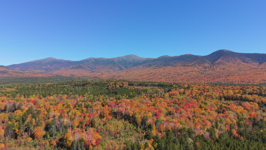 Rolling spruce ridge rises toward Mount Washington, autumn-colored valley floor stretching beneath layered summits across White Mountain National Forest in New Hampshire. Aerial drone rising wide shot