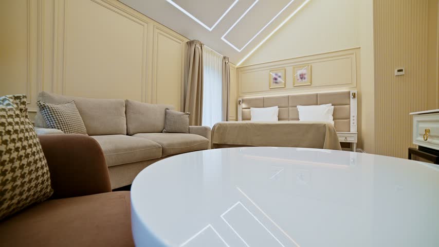 A modern luxury hotel room interior features a sitting area and a bed. The camera pans smoothly to the right across the room. The space includes wooden flooring and warm beige walls.