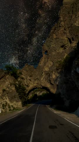 Milky Way Galaxy view above an asphalt road passing through a natural rock arch in the mountains at night. Road through the cave