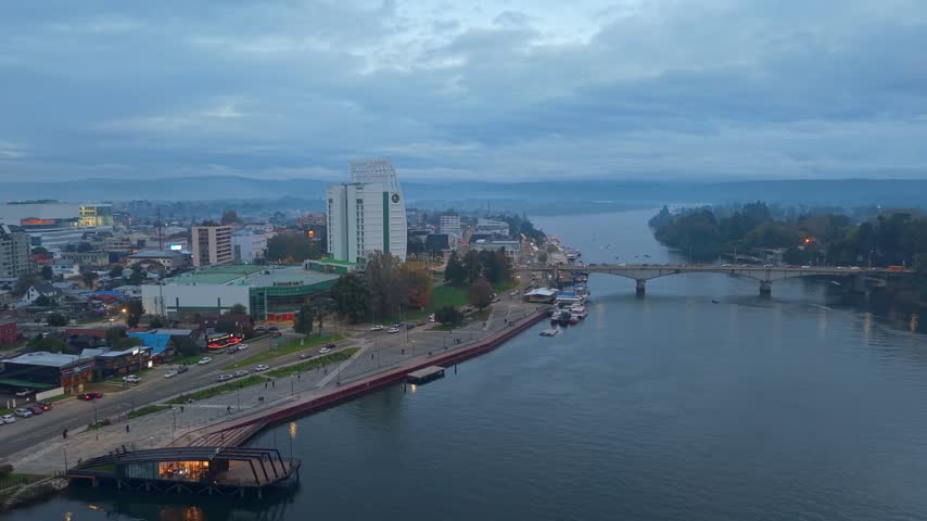Aerial drone footage capturing cityscape of Valdivia Chile during twilight with bridge traffic and calm river waters flow.