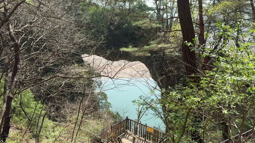 Scenic view looking through trees at massive natural rock formation shaped like bridge over turquoise river water in serene forest setting.