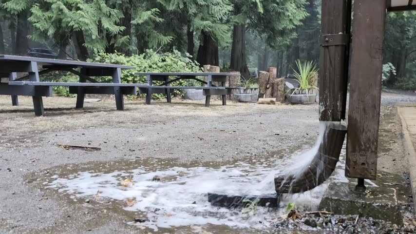 Heavy Rainwater Pouring From Wooden Gutter Downspout Onto Ground Pool Beside Picnic Tables In Forest Park. - closeup shot