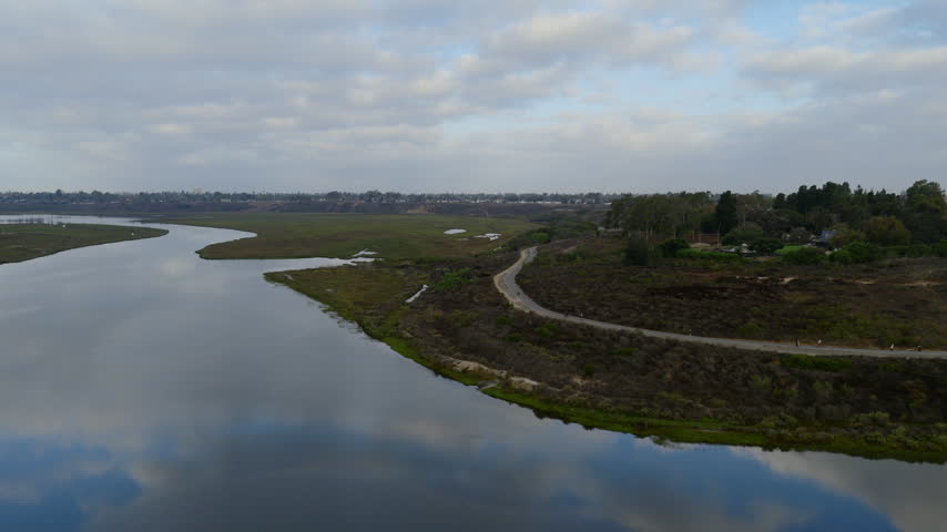 Aerial view of a winding trail along the scrub-covered shore of Upper Newport Bay, with walkers on the path, calm water reflecting clouds, and salt marsh stretching to the horizon.