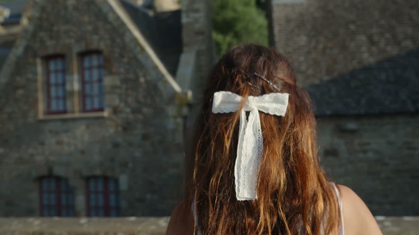 Back view of a woman with long brown hair and a white ribbon looking toward Mont Saint Michel Abbey above medieval stone rooftops in France