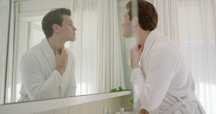 Adult man in bathrobe leaning toward mirror, inspecting and massaging jawline at vanity, copy space. Grooming, skincare, selfcare, reflection, bathroom, softlight, countertop
