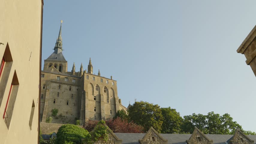 Mont Saint Michel Abbey towers above historic stone houses and slate rooftops in France, framed by green trees under a clear blue sky