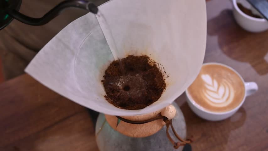 Barista making pour over coffee next to a cup of cappuccino with latte art on a wooden table
