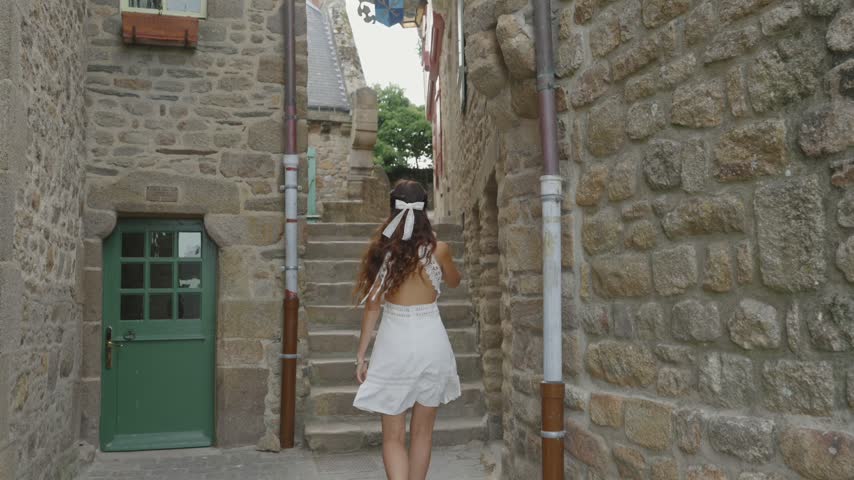 Back view of a woman in a white dress climbing narrow medieval stone steps between historic buildings at Mont Saint Michel, France