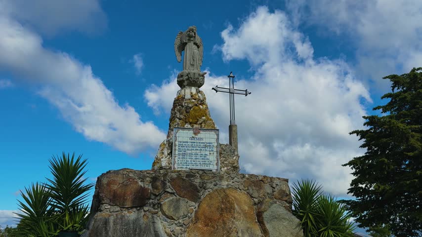 A historic stone angel sculpture and a metal cross standing atop a large rock monument under a vibrant, cloudy blue sky.
