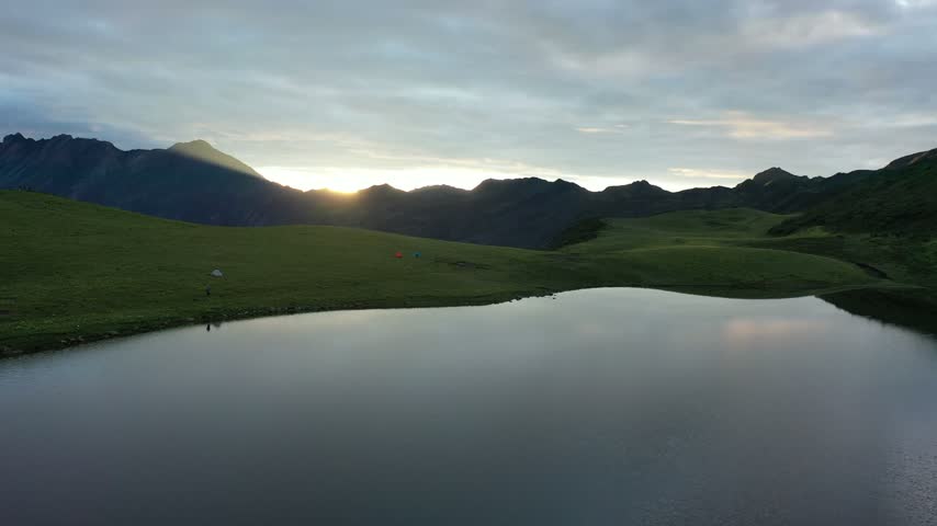 Aerial drone footage at sunrise over Jiudingshan Heilongchi near Chengdu, China, showing green alpine meadows, flower fields, a calm mountain lake and silhouetted peaks with golden morning light.