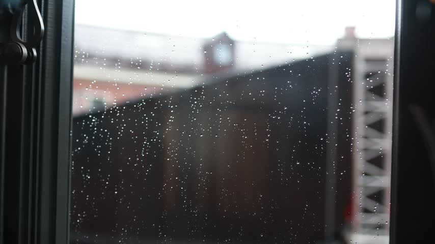 Close-up of raindrops on window glass with a black latch and blurred buildings outside on a gray day