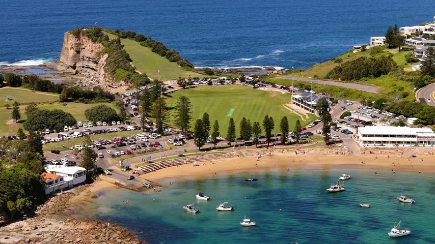 Coastal park and sandy bay meet the rocky Terrigal headland beside clear blue water and waterfront development, aerial