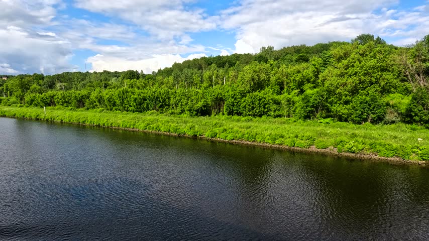 A white train moves through a dense green forest along a calm river under a partly cloudy sky. The still water and lush trees create a peaceful, serene landscape.
