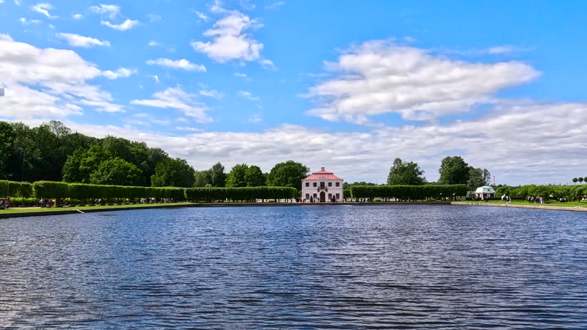 A peaceful park scene with a white building located above a large lake surrounded by neat hedges and trees against a blue sky background. People are strolling along the paths, enjoying the scenery.
