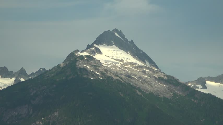 Close-up view of a rugged, snow-capped mountain peak within the Tantalus Mountain Range near Squamish, British Columbia. The glacier-carved alpine summit towers under a hazy blue summer sky.