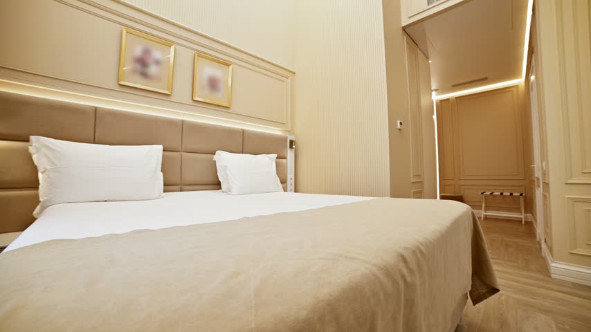 The camera moves slowly forward through a modern luxury hotel room interior. The low-angle shot features a large bed, textured walls, and warm linear lighting.