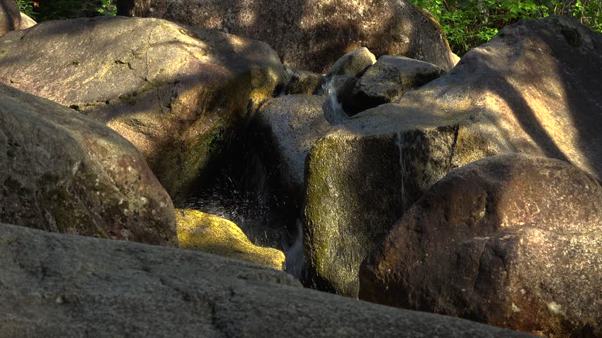 Close-up of fresh water trickling and splashing between massive sunlit granite boulders at Shannon Falls in Squamish, British Columbia. The dark stone rocks create a textured abstract gap.