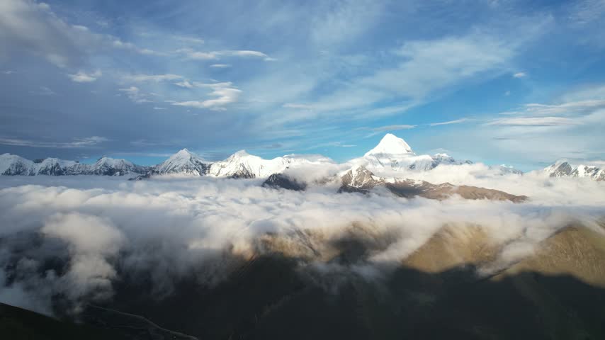 Aerial drone footage of Mount Gongga in Sichuan, China, with snow-capped peaks rising above a vast sea of clouds and flowing cloud waterfalls under a clear blue sky.