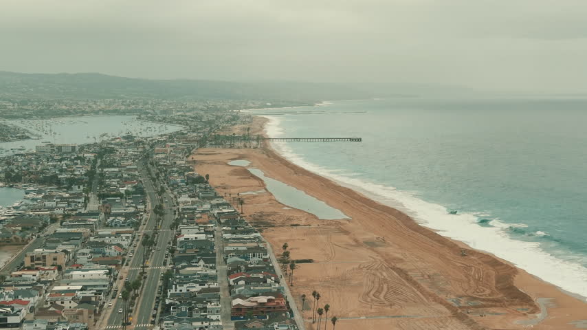 Drone panorama of Newport Beach coastline showing ocean waves, sand barriers and coastal homes.