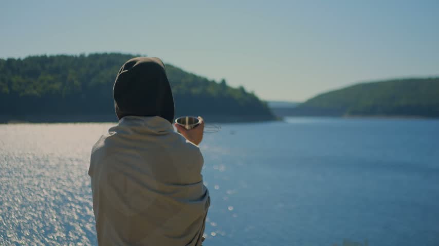 Woman holding a cup beside the calm waters of Colebrook River Lake, CT, surrounded by forested hills, conveying mindfulness, peace, relaxation, and connection with nature. 4K.
